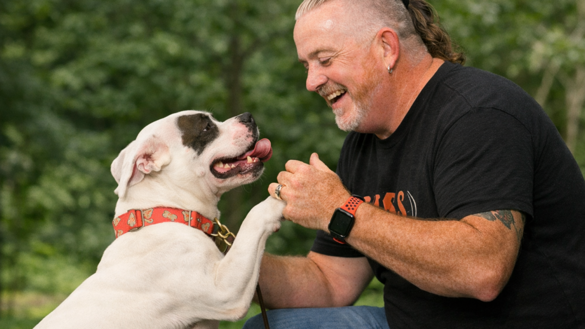 Dog trainer Mike Deathe kneeling and playfully engaging with a dog during an outdoor training session, illustrating how fun, consistency, and management improve training results.