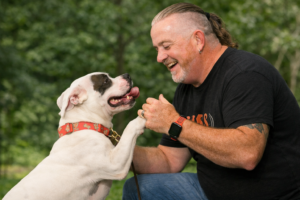 Dog trainer Mike Deathe kneeling and playfully engaging with a dog during an outdoor training session, illustrating how fun, consistency, and management improve training results.